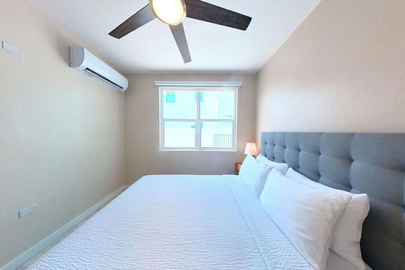 Une chambre à coucher comprenant un lit, un ventilateur de plafond, un appareil de climatisation, une fenêtre et une table de chevet avec une lampe.