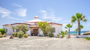 A white building with a dome and red roof, surrounded by gravel and palm trees, with a view of the ocean.