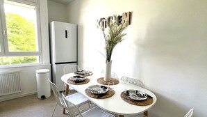 Un moderno comedor con una mesa blanca dispuesta para cuatro personas, rodeada de sillas blancas. Una lámpara decorativa de metal cuelga del techo y se ve un radiador blanco en la pared.