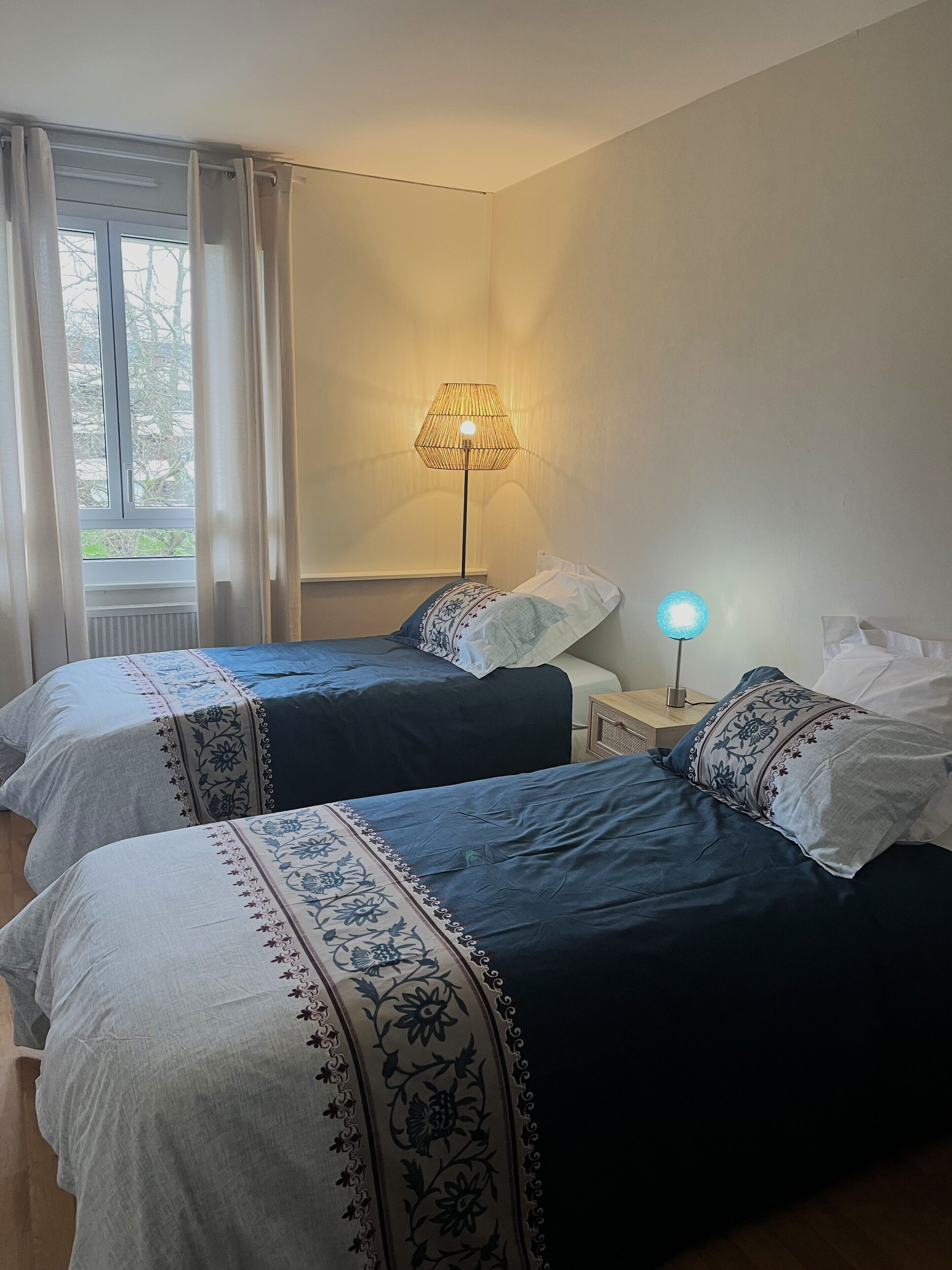 Una stanza con due letti, un comodino con lampada e una finestra con tende.
