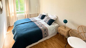 Un dormitorio con una cama grande, una mesita de noche con lámpara, una silla y vistas al verdor exterior.