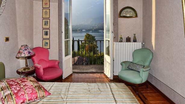 A room with a bed, two chairs, a lamp, and a view of a lake and mountains through an open door. - Villa Emilia Apartment by Wonderful Italy (Bellagio)
