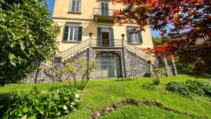 A two-story building with a stone base and a yellow facade, featuring a balcony and a large window at the ground level. - Villa Emilia Apartment by Wonderful Italy (Bellagio)