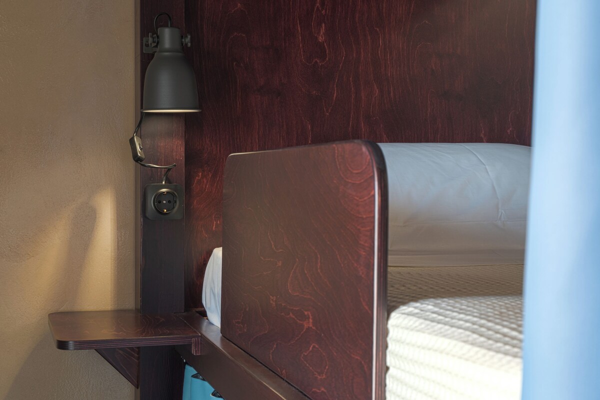 Une tête de lit en bois, un matelas blanc, une lampe fixée au mur et une prise de courant.