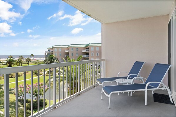 A balcony with two lounge chairs and a small table, overlooking a landscaped area with palm trees and a view of buildings in the distance. - St. Simons Grand 214 (St Simons Island)