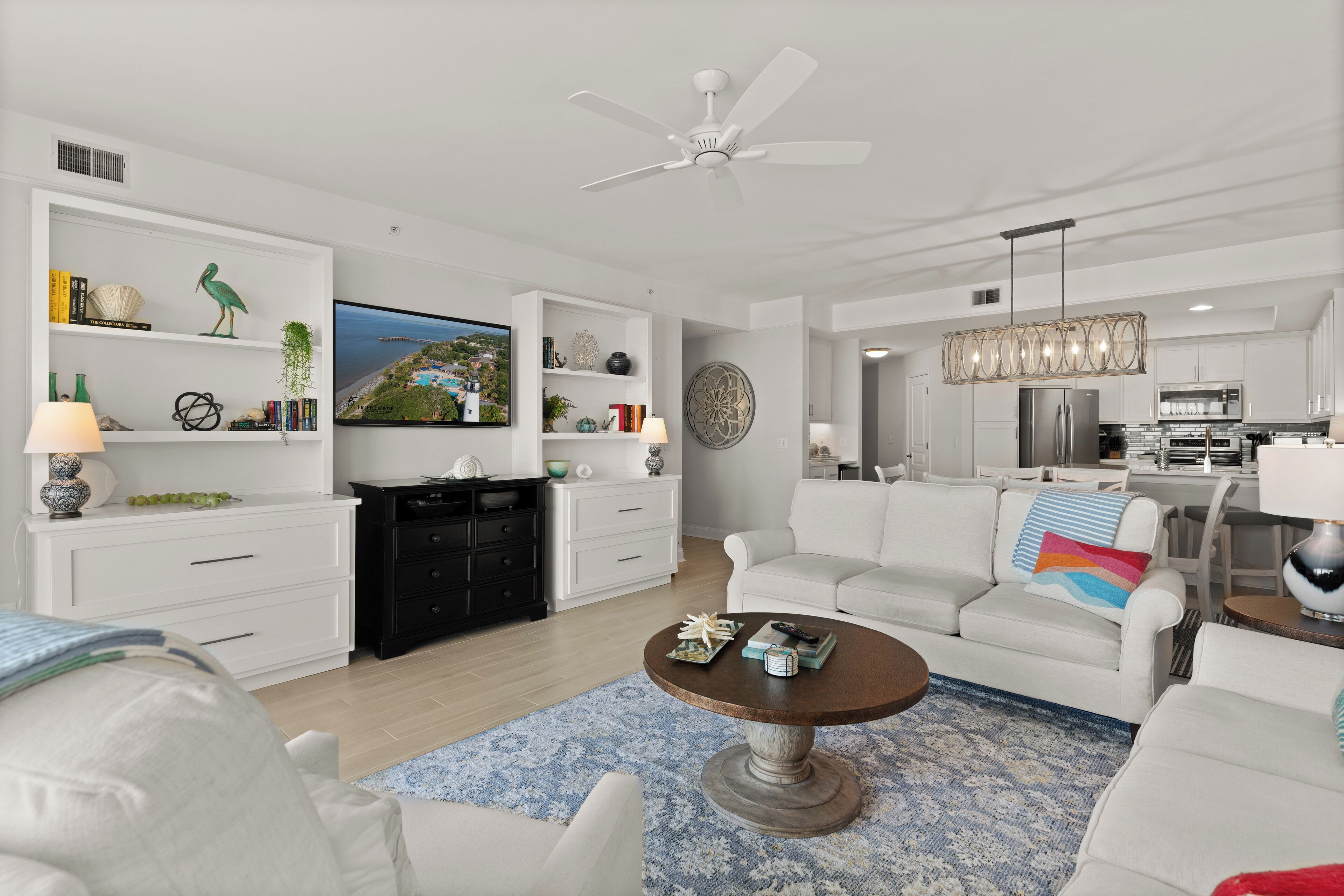 Una sala de estar moderna con muebles blancos, un televisor y una cocina con gabinetes blancos y un refrigerador de acero inoxidable.