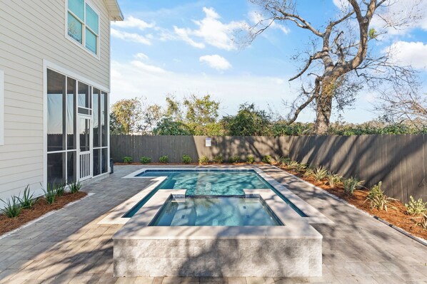 A rectangular swimming pool with a stone border, surrounded by a paved patio with a wooden fence and a tree in the background. - Time Fly’s when at the Beach, sleeps 8 (St Simons Island)