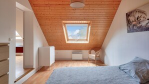 Un dormitorio con techo de madera inclinado, un tragaluz, una cama, una silla y un radiador.