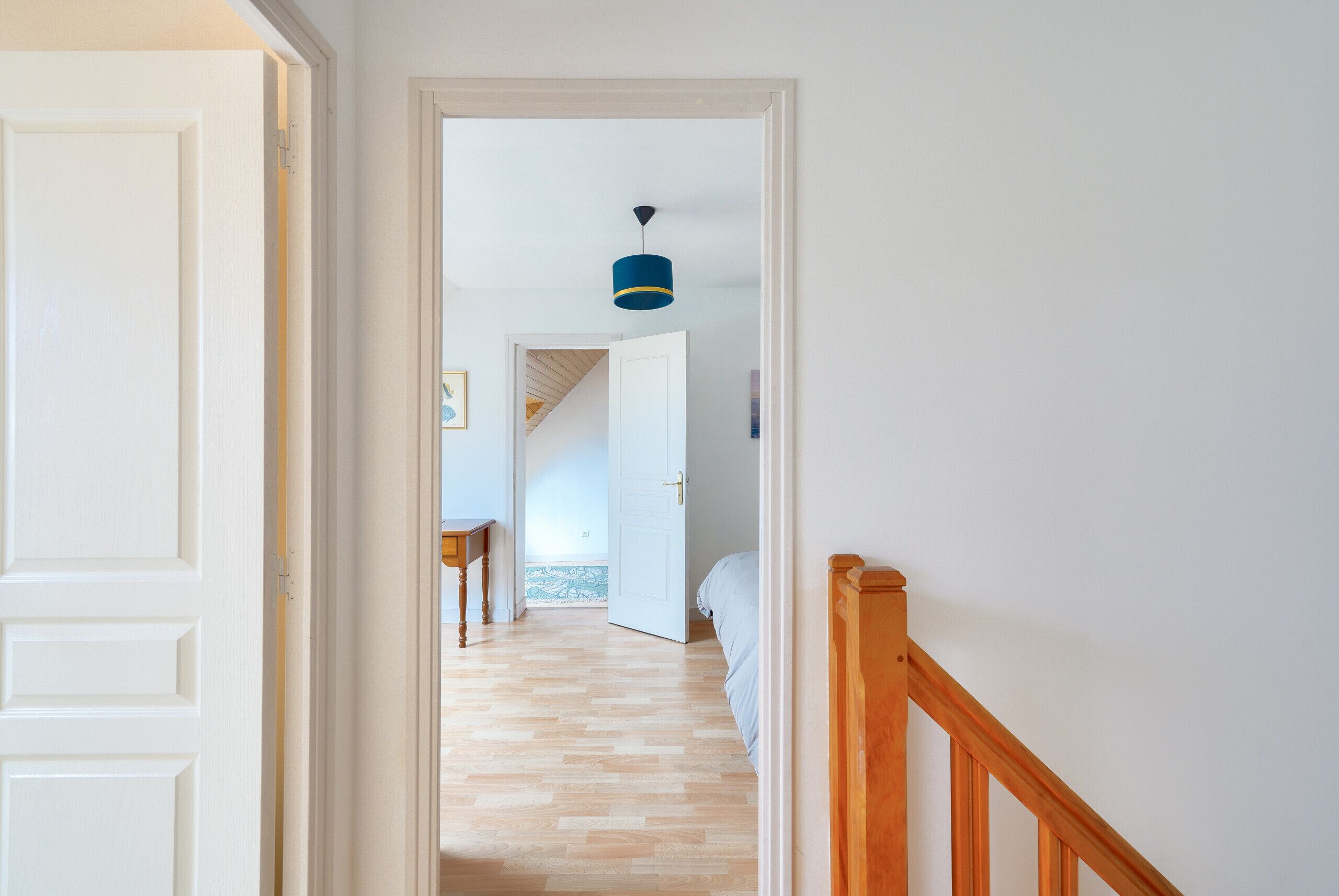 En hall som leder till ett sovrum med trägolv, en säng och en blå hänglampa.
