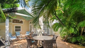 A wooden deck with wicker furniture and a round table, surrounded by lush tropical plants. - Find your zen at HIDDEN TREASURE (Key West)