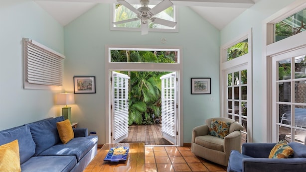 A living room with a blue sofa, a beige armchair, and a wooden coffee table. There is a ceiling fan, a window with blinds, and a door leading to a garden. - Find your zen at HIDDEN TREASURE (Key West)
