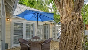 A covered patio with wicker furniture and a blue umbrella, surrounded by a large tree and a white fence. - Get your ISLAND STLYE on! (Key West)