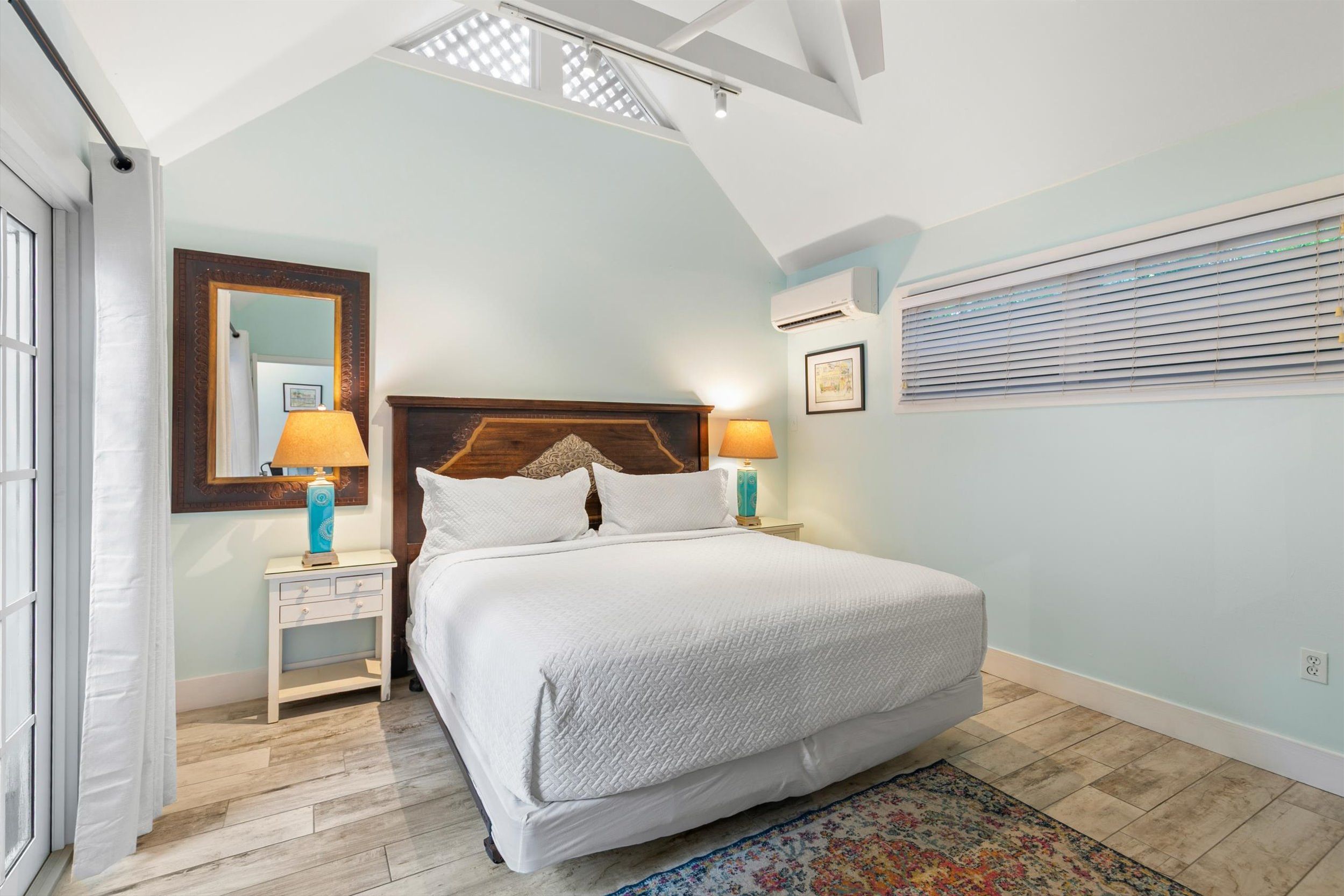 A bedroom with a bed, bedside tables, a mirror, an air conditioning unit, and a window with blinds.