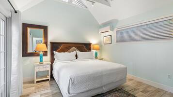 A bedroom with a bed, bedside tables, a mirror, an air conditioning unit, and a window with blinds.