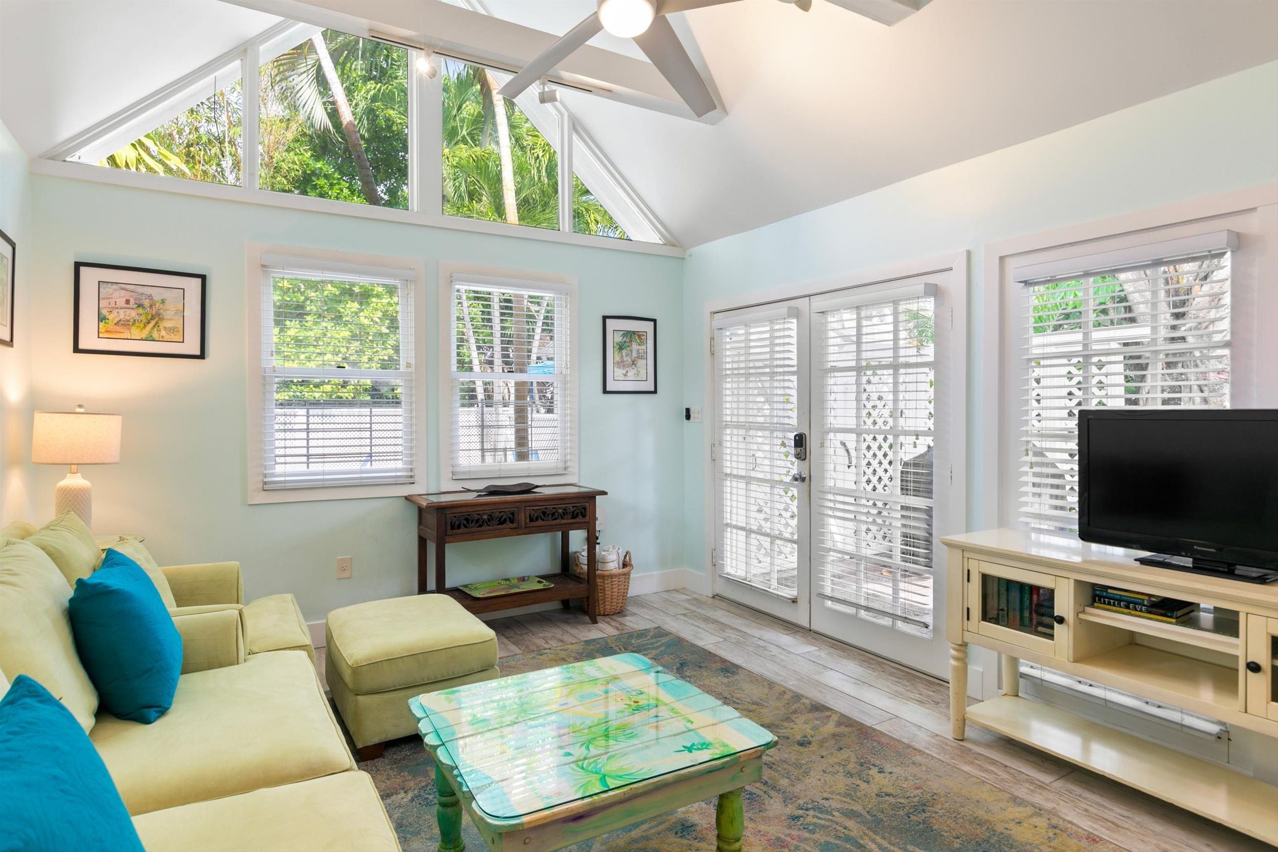 A living room with a sofa, ottoman, coffee table, TV stand, and large windows with blinds.