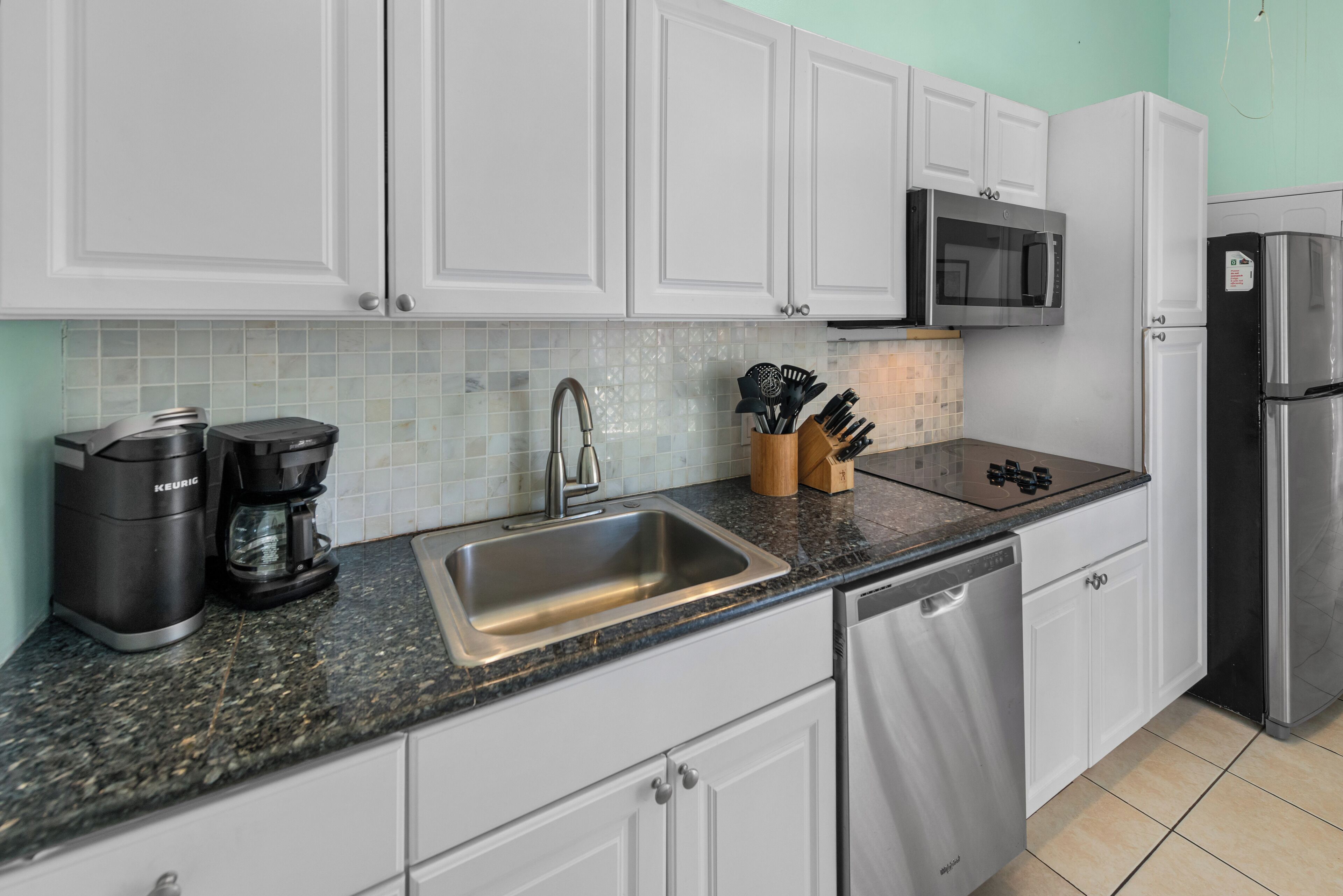 A modern kitchen with white cabinets, a stainless steel sink, a coffee maker, a microwave, and a refrigerator.