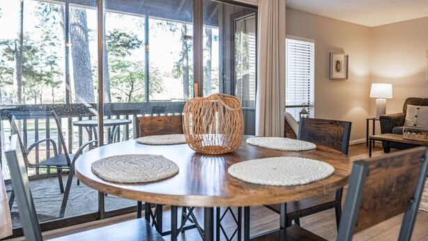 A dining area with a round table, chairs, a woven basket, and a view of trees through large windows.