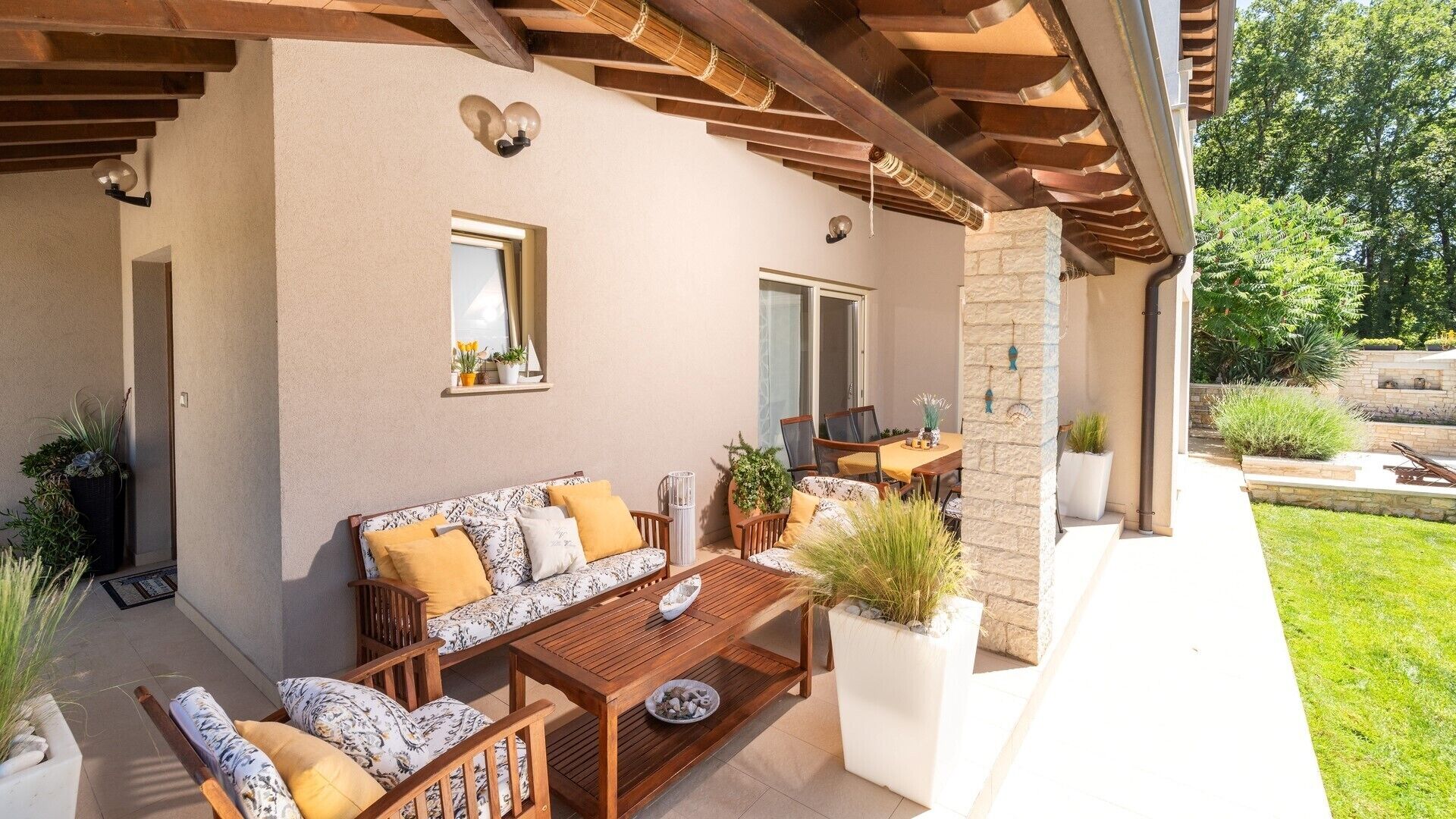 Eine Terrasse im Freien mit einer Holzbank, einem Tisch und einer Couch. Es gibt Kübelpflanzen und eine Steinmauer.