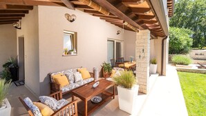 An outdoor patio with a wooden bench, a table, and a sofa. There are potted plants and a stone wall.