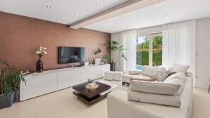 A modern living room with a flat-screen TV, a sofa, a coffee table, and a large window with a view of greenery.