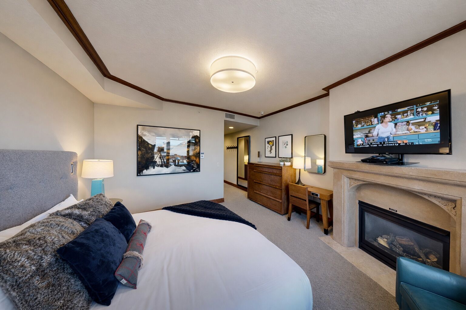 A hotel room with a large bed, a desk, a television, and a fireplace.