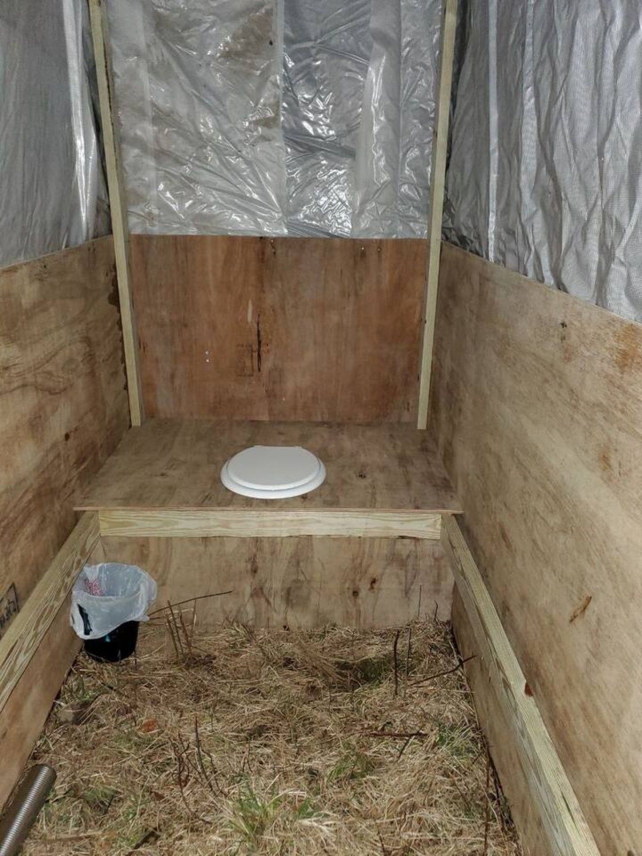 A small, enclosed wooden structure with a white circular object on the floor, possibly a toilet seat, and a plastic bag on the ground.