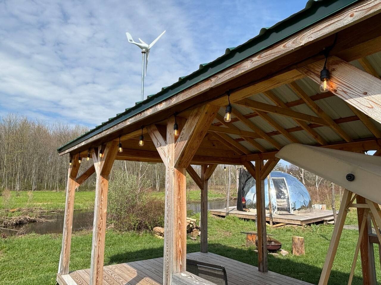 A wooden pavilion with a metal roof, a wind turbine in the background, and a covered vehicle in the distance.