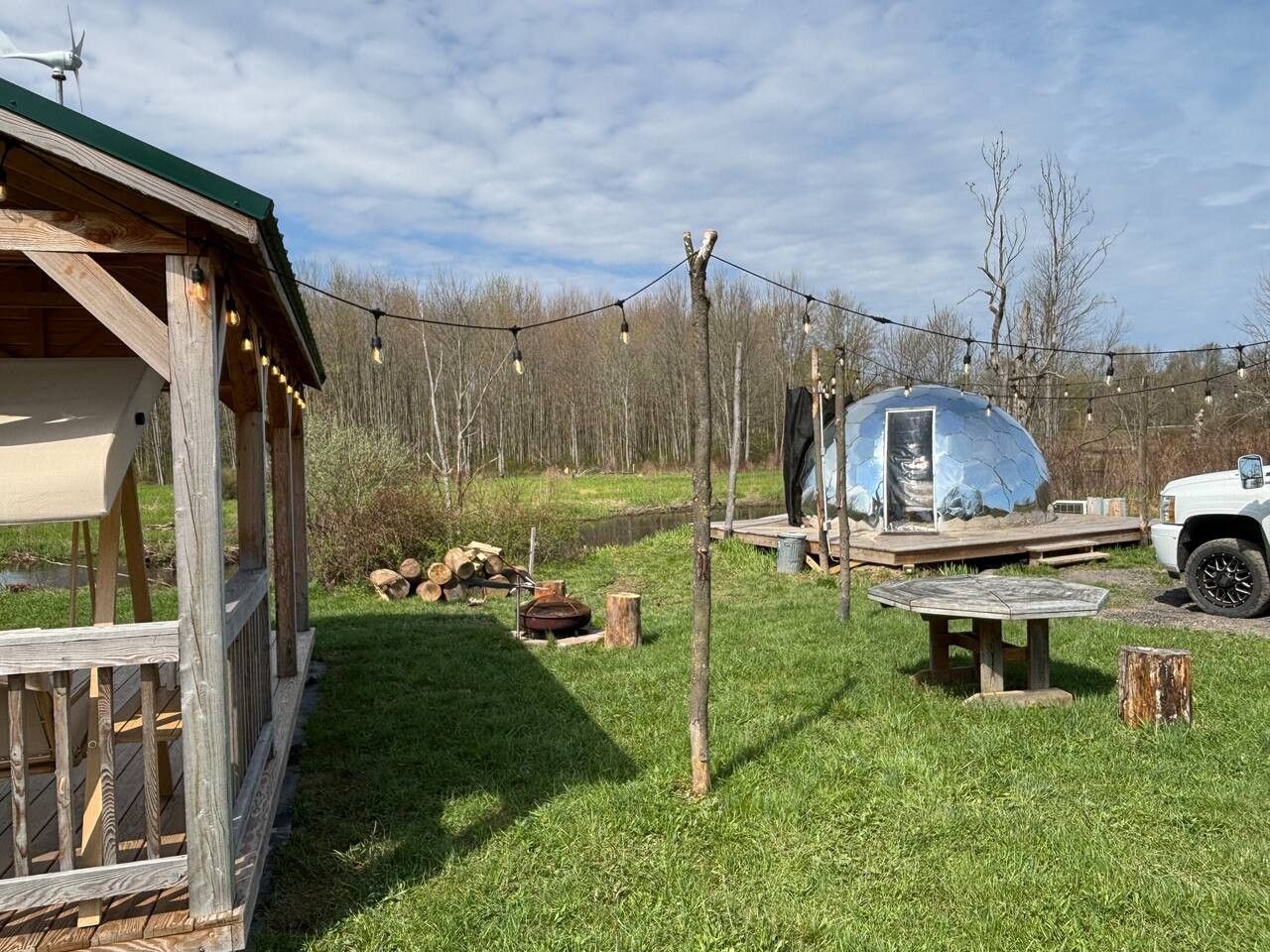 A rustic outdoor area with a wooden deck, a covered seating area, a fire pit, and a large spherical structure.