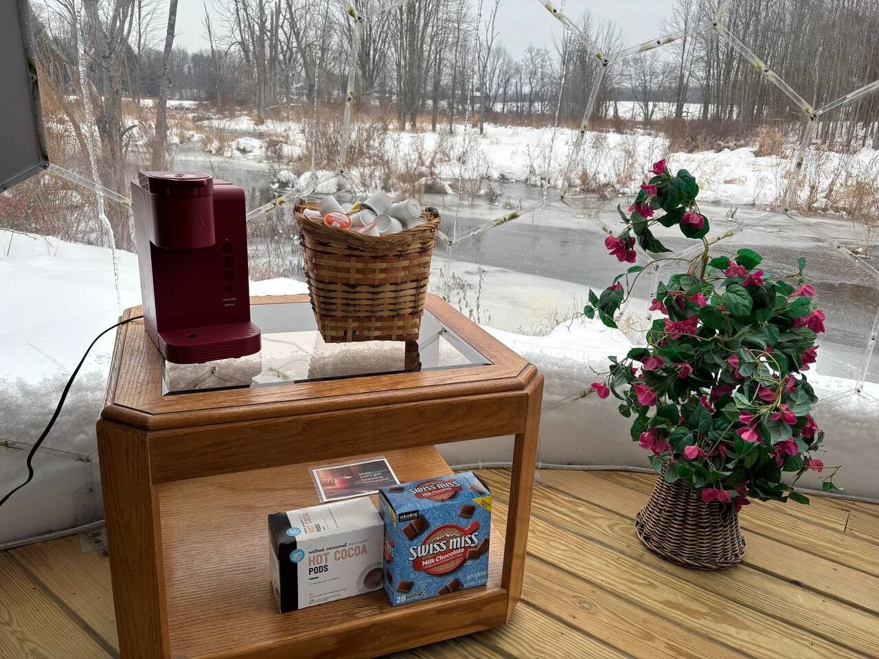 Une table en bois, sur laquelle se trouvent une cafetière rouge, un panier rempli de tasses et une boîte de dosettes de chocolat chaud Swiss Miss.