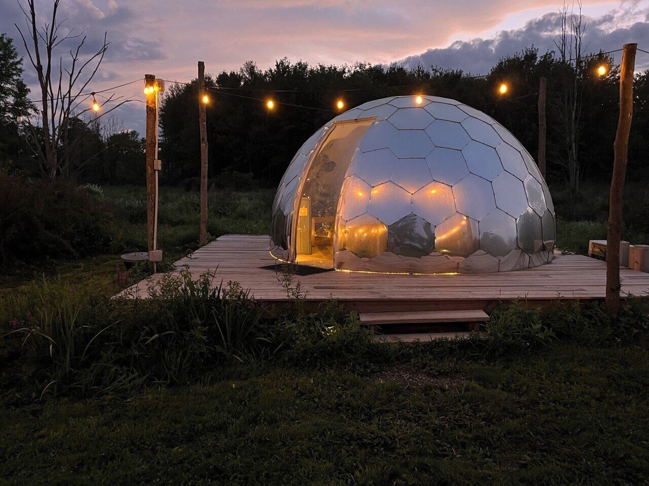 A geodesic dome structure with a wooden deck, surrounded by trees and illuminated by string lights.