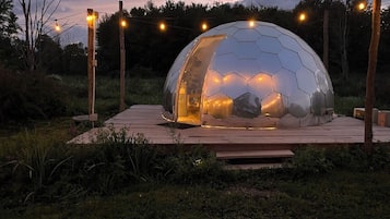 A geodesic dome structure with a wooden deck, surrounded by trees and illuminated by string lights.