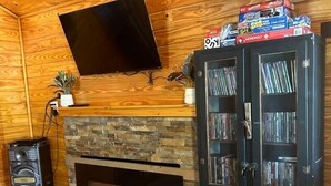 Uma sala de estar com lareira de pedra, paredes de madeira, uma televisão de tela plana fixada acima e uma estante cheia de DVDs.