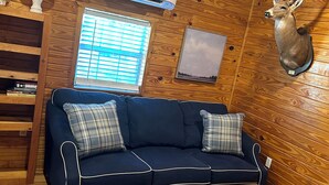 Um quarto aconchegante com um sofá azul-escuro, uma mesa de centro de madeira, uma luminária, uma decoração de cabeça de veado, uma janela com persianas e uma estante de livros.