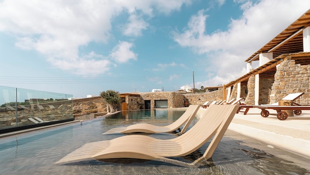 A clear swimming pool with lounge chairs, stone walls, and a modern building with a sloped roof. - MvibesMykonos homes and studios (Mykonos)