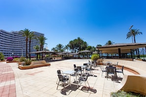 An outdoor patio area with tables and chairs, surrounded by palm trees and a large building in the background.