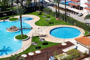 A large outdoor swimming pool with multiple sections, surrounded by palm trees, umbrellas, and a well-maintained lawn.
