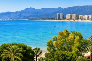 A coastal cityscape with a beach, clear blue waters, and mountains in the background.
