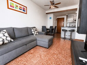 Una sala moderna con un sofá seccional gris, un pequeño reposapiés, un ventilador de techo, un comedor con mesa y sillas, y una zona de cocina con barra.