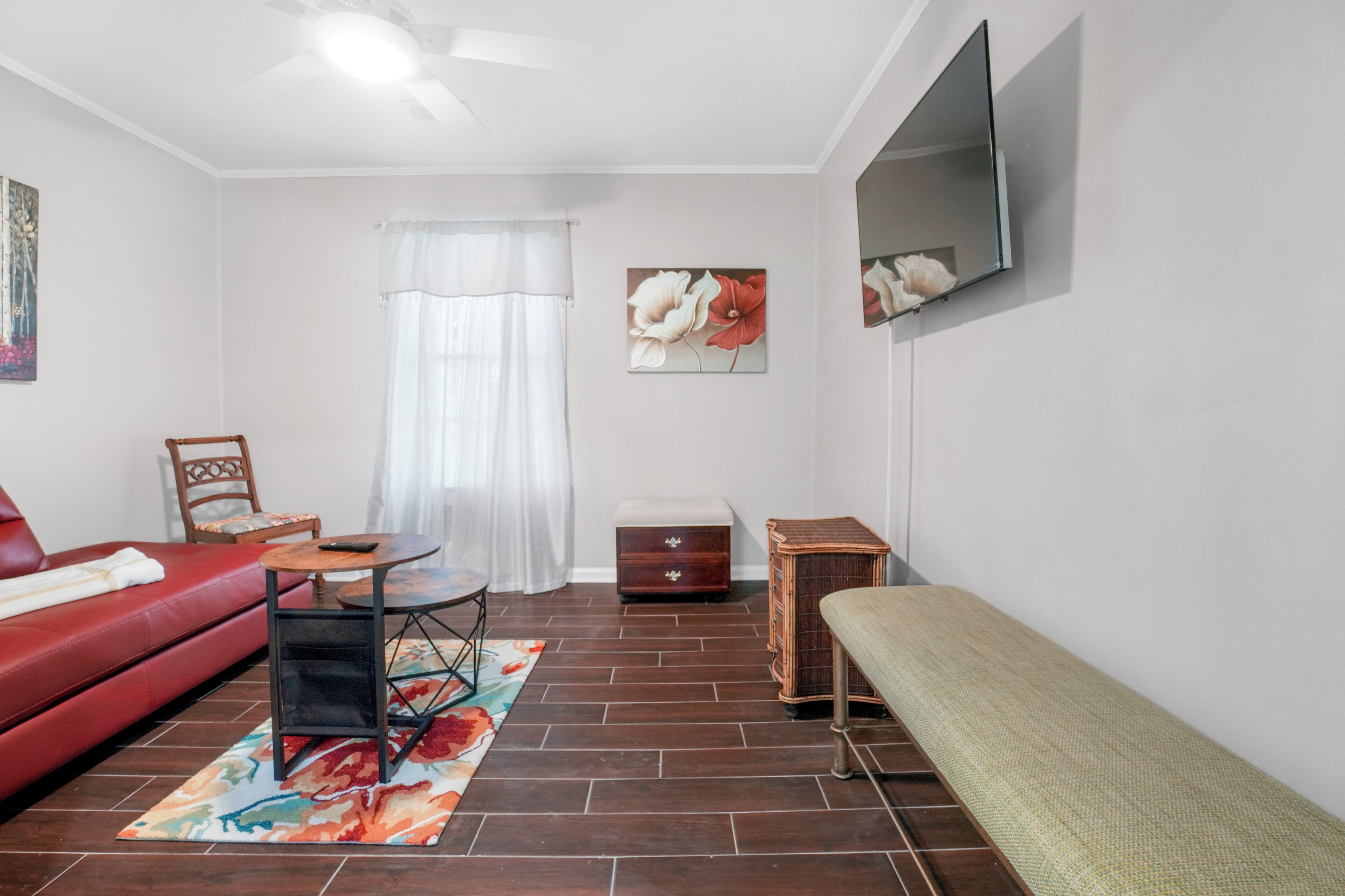 Un salon avec un canapé rouge, un banc en bois, une petite table, un téléviseur fixé au mur et une fenêtre ornée de rideaux blancs.