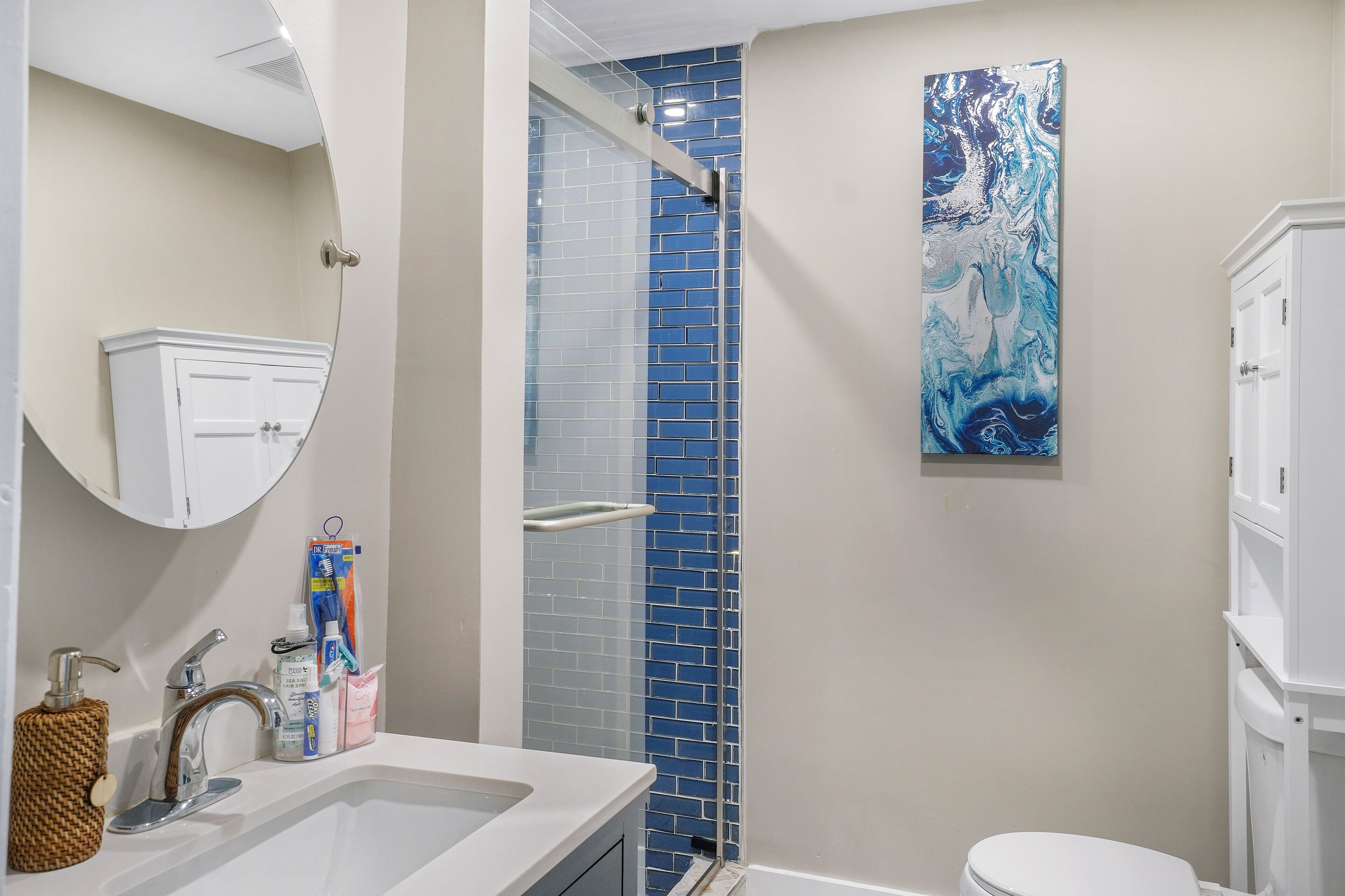 A bathroom with a white sink, a mirror, a white cabinet, a blue-tiled shower enclosure, and a large abstract painting on the wall.