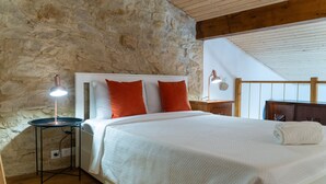 A bedroom with a stone wall, wooden ceiling beams, a bed with white bedding and orange pillows, a bedside table with a lamp, and a wooden nightstand. - Casa S o Jos in Carvalhal (Bombarral)