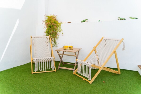 Twee houten schommelstoelen met witte netten en kwastjes, een klein houten tafeltje met een gele kom, en een potplant voor een witte muur.