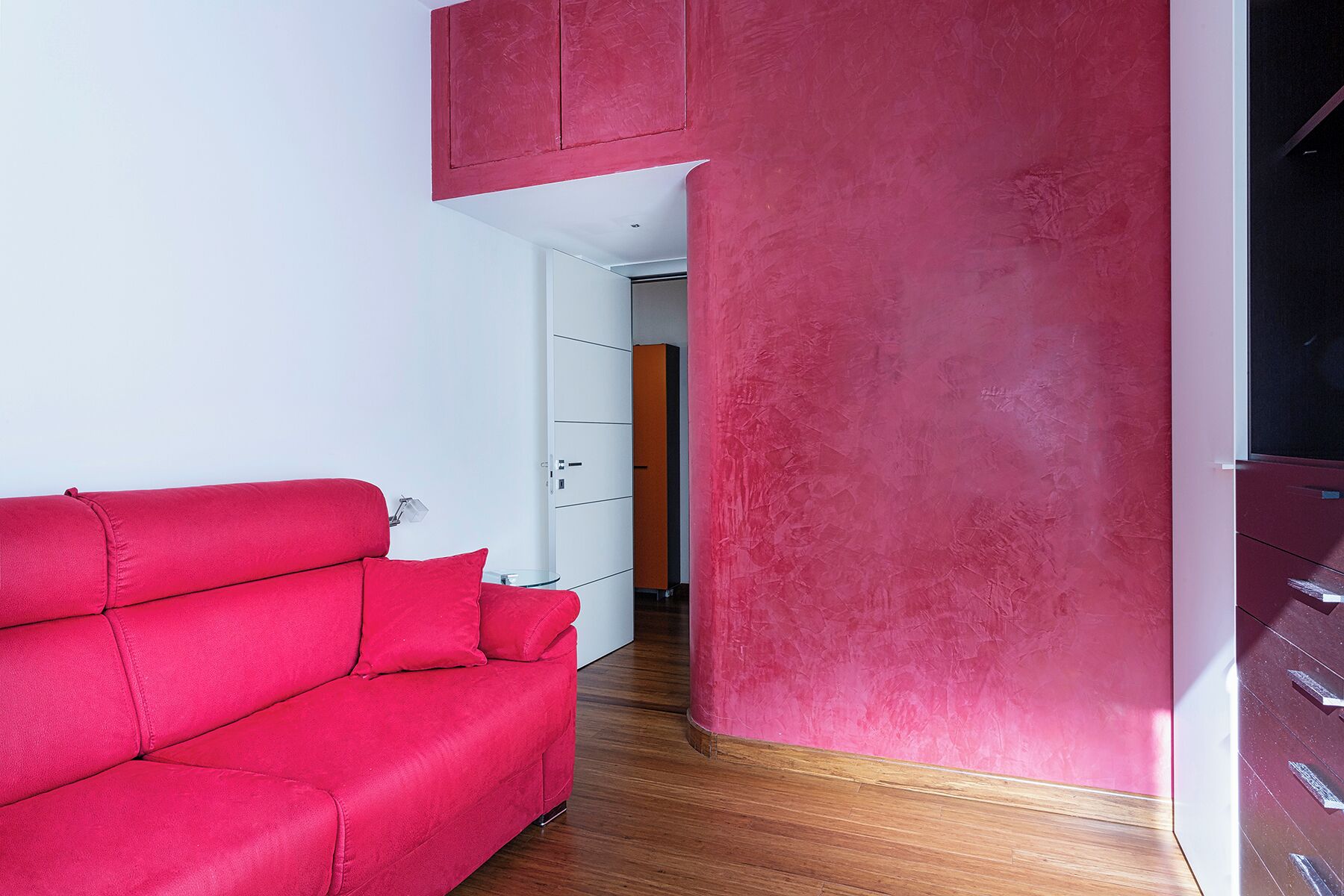 A modern living room with a red sofa, wooden flooring, and a pink accent wall.