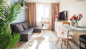 Moderni olohuone, jossa on harmaa sohva, ruokapöytä tuoleineen, televisio ja suuri huonekasvi.