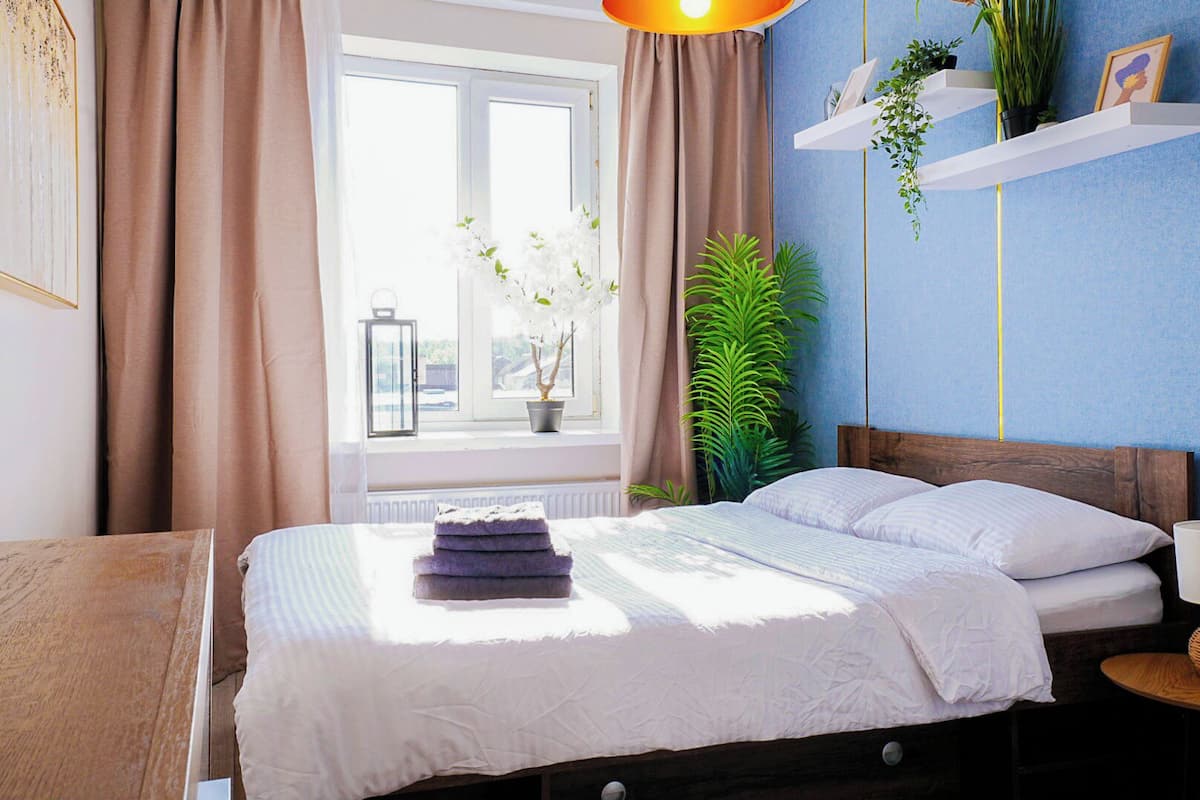 Un lit bien fait, recouvert d’une couette blanche, et une pile de serviettes pliées. Il y a une fenêtre avec des rideaux, une plante en pot et une étagère fixée au mur, sur laquelle se trouvent un cadre photo et une petite plante.