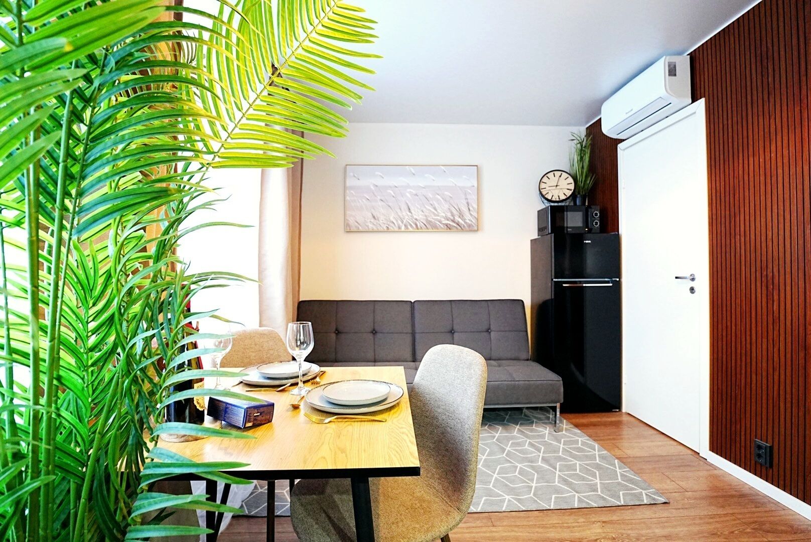 A dining area with a table set for two, a sofa, a clock, and a microwave.