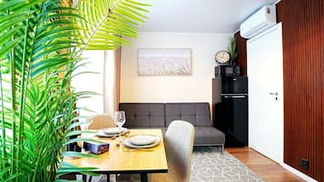 A dining area with a table set for two, a sofa, a clock, and a microwave.