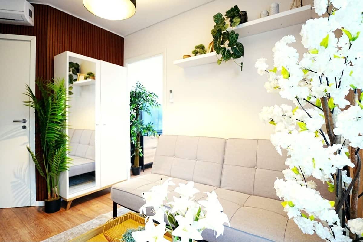Un salon moderne avec un canapé beige, un plancher en bois et diverses plantes, dont une grande plante verte et des fleurs blanches.