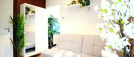 Una sala de estar moderna con un sofá color crema, piso de madera y varias plantas, incluyendo una planta verde alta y flores blancas.