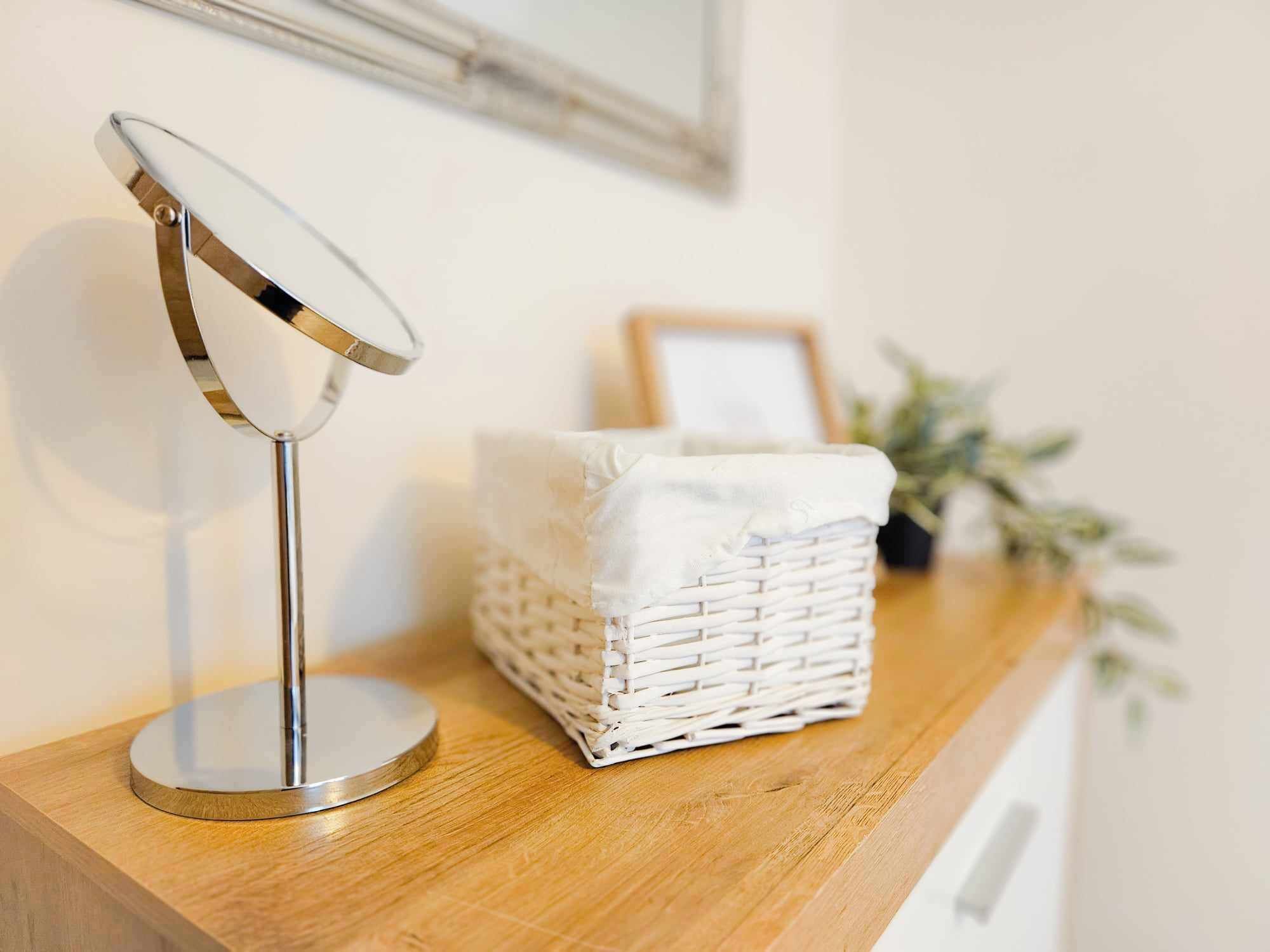 A round mirror on a wooden surface, a white wicker basket, and a framed picture on the wall.